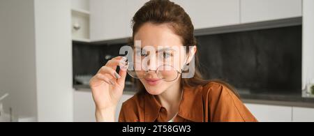 Porträt einer schönen jungen Frau, lächelnd und glücklich aussehend, zieht ihre Brille ab und blickt in die Kamera, sitzt zu Hause in der Küche Stockfoto