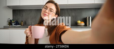 Porträt einer schönen jungen Frau in Brille, macht Selfie mit einer Tasse Tee, sitzt in der Küche, hält Smartphone Stockfoto