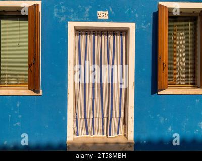 Ein Haus in Burano in Venedig, Italien. Ein blau-weiß gestreiftes Blatt verbirgt den Eingang. Zwei Fenster mit Holzläden und grünen Jalousien runden das ab Stockfoto