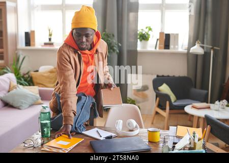 Horizontale Aufnahme eines modernen afroamerikanischen College-Studenten, der am Schreibtisch steht und sich auf die Schule vorbereitet, indem er Lehrbücher und Copybooks in den Rucksack packt Stockfoto