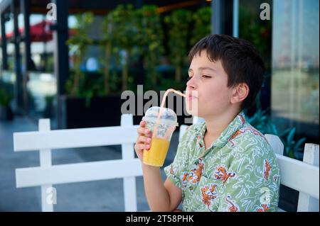 Seitenporträt eines hübschen kaukasischen dunkelhaarigen Teenagers in Sommerkleidung, der frisch gepressten Saft aus einem Strohhalm trinkt und ca. Stockfoto