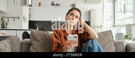 Porträt einer niedlichen neugierigen Frau, die mit dem Smartphone auf dem Sofa sitzt, aufblickt und nachdenkt, eine Bestellung aufgibt, Online-Shopping über die Handy-App Stockfoto