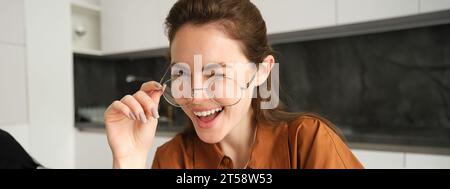 Porträt einer schönen jungen Frau, lächelnd und glücklich aussehend, zieht ihre Brille ab und blickt in die Kamera, sitzt zu Hause in der Küche Stockfoto