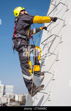 Ein männlicher Industriekletterer klettert in Sicherheitsausrüstung und Helm auf eine Metallleiter, um Montagearbeiten durchzuführen. Durchführung von Hochhausarbeiten an Metallkonstruktionen. Stockfoto