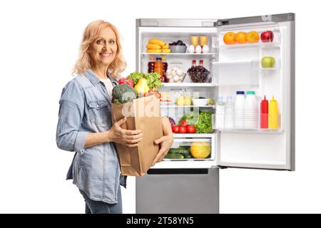 Reife Frau mit einem Lebensmittelbeutel neben einem offenen Kühlschrank mit Essen isoliert auf weißem Hintergrund Stockfoto