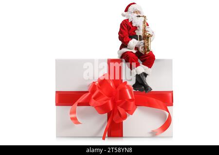 Weihnachtsmann spielt Saxophon und sitzt auf einer großen Geschenkbox mit roter Schleife isoliert auf weißem Hintergrund Stockfoto