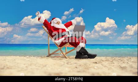 Santa claus am Strand, der auf einem Liegestuhl mit Laptop sitzt und Glücksgefühle ausdrückt Stockfoto