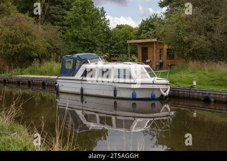 Ein schmales Boot liegt auf einem Kanal vor einem Holzschuppen. Es spiegelt sich perfekt im stillen Wasser wider Stockfoto