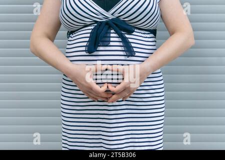 Schwangere Frau mit Herzzeichen auf dem Bauch. Schwangere Frau, die auf gestreiftem Hintergrund steht Stockfoto