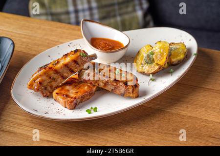 Gegrillte Schweinerippchen serviert mit gebratenen neuen Kartoffeln auf einem weißen Teller auf einem Holztisch. Gesundes Essen, Nahaufnahme. Serviert Speisen in einem Restaurant Stockfoto
