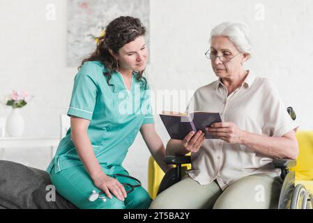 Behinderte Seniorin sitzender Rollstuhl-Lesebuch mit Krankenschwester Stockfoto