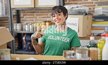 Junge, schöne hispanische Frau, die auf dem Tisch sitzt und auf freiwillige Uniform zeigt, lächelt im Wohltätigkeitszentrum Stockfoto