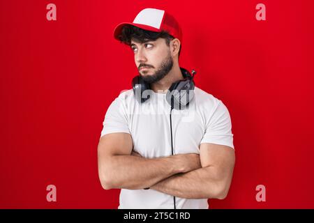 Hispanischer Mann mit Bart, Gamer-Hut und Kopfhörer, der mit überkreuzten Armen zur Seite blickt, überzeugt und selbstbewusst Stockfoto