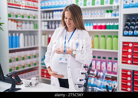 Junge schwangere Apothekerin, die selbstbewusst lächelt und den Bauch in der Apotheke berührt Stockfoto