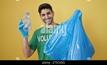 Junger hispanischer Freiwilliger, der Handschuhe trägt und Plastikflaschen auf isoliertem gelbem Hintergrund recycelt Stockfoto