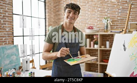 Selbstbewusster junger hispanischer Teenager-Künstler mischt fröhlich die Farbpalette und lächelt beim Basteln im Kunststudio Stockfoto