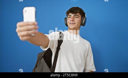 Cooler junger hispanischer Teenager, ein lächelnder Student, der in ein lebhaftes Videogespräch vertieft ist, isoliert vor einem blauen Hintergrund steht Stockfoto