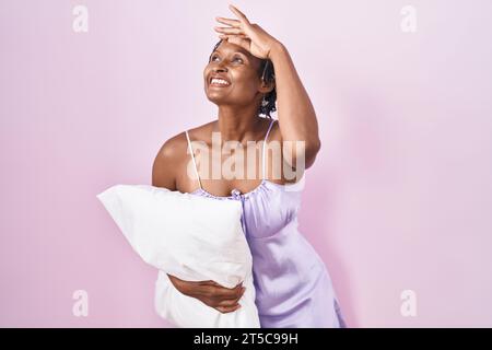Afrikanische Frau mit Dreadlocks, die Pyjama trägt und das Kissen umarmt, sehr glücklich und lächelnd, mit Hand über Kopf weit weg. Suchkonzept. Stockfoto