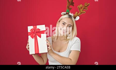 Junge schöne hispanische Frau trägt Rentier weihnachtshut mit Geschenk über isoliertem roten Hintergrund Stockfoto