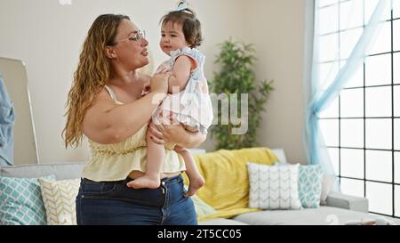 Glückliche Mutter steht zuversichtlich zu Hause, hält ihre liebenswerte Tochter in den Armen und genießt gemeinsam die Liebe der Familie in ihrem Wohnzimmer mit einem Stockfoto