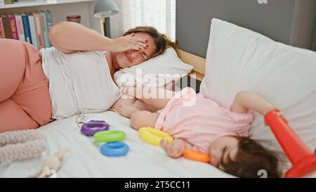 Gestresste Mutter und Tochter liegen im Bett, während Baby mit Spielzeug im Schlafzimmer spielt - ein ergreifender Familienmoment Stockfoto
