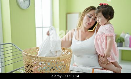 Die schöne Mutter hält ihre kleine Tochter in den Armen, spricht mit dem Smartphone, während sie die Kleidung gemütlich an der Wäscheleine hängt und die lässige Arbeit in ho genießt Stockfoto