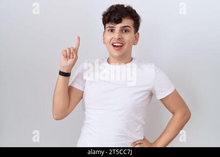 Junger, nicht binärer Mann mit lässigem weißem T-Shirt, der den Finger mit einer erfolgreichen Idee nach oben zeigt. Verlassen und glücklich. Nummer eins. Stockfoto