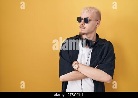Junger kaukasischer Mann mit Sonnenbrille, der über gelbem Hintergrund steht und mit überkreuzten Armen zur Seite schaut Stockfoto