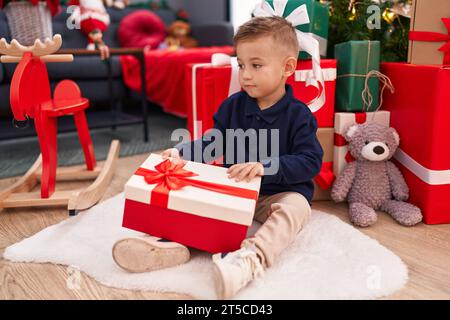 Entzückender hispanischer Junge, der zu Hause am weihnachtsbaum sitzt und ein Geschenk auspackt Stockfoto