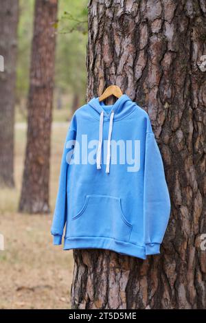 Blauer Kapuzenpullover, der an einem Bügel an einem Baum im Wald hängt. Darstellung verschiedener Farben der Kleidung Stockfoto