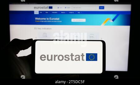 Person mit Mobiltelefon mit Logo des EU-Organs Europäisches Statistisches Amt Eurostat vor der Unternehmenswebseite. Schwerpunkt auf dem Display des Telefons. Stockfoto