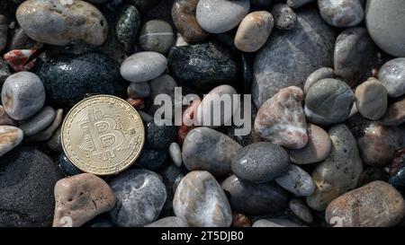 Goldene Bitcoin-Münze vor einem Hintergrund großer grauer Steine entlang der Küste, die die zeitgenössische Stabilität der elektronischen Währung symbolisieren. Stockfoto