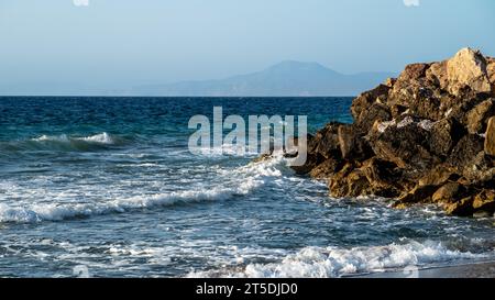 Faszinierende Landschaft mit dem Meer, in dem Wellen rauschen und gegen Steinbrecher krachen, mit Bergen, die in der Ferne sichtbar sind. Stockfoto