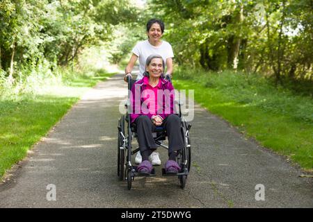 Asiatische Indianerin schubst ihre ältere Mutter im Rollstuhl im Sommer in Großbritannien nach draußen. Stellen Sie möglicherweise auch einen Betreuer, Fürsorge in der Gemeinde dar Stockfoto