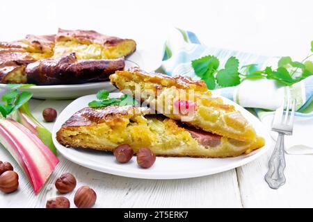 Zwei Stücke Torte mit Rhabarber und Nusscreme auf einem Teller, Rhabarberstiele und Nüsse, Serviette und Minze auf Holzbrett Hintergrund Stockfoto