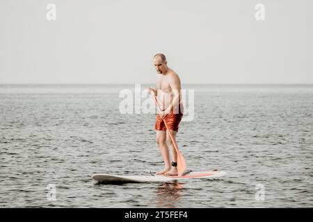 Aktiver reifer männlicher Paddler mit seinem Paddleboard und paddeln im Sommer auf dem Meer. Ein glücklicher Seniorenmann steht mit einem SUP-Brett. Stand-Up-Paddle-Boarding - Stockfoto