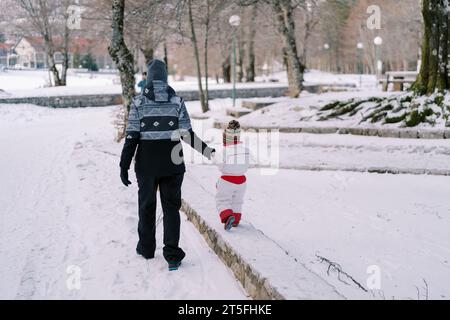 Mutter führt ein kleines Kind an der Hand entlang der schneebedeckten Bordsteinkante im Park. Rückansicht Stockfoto