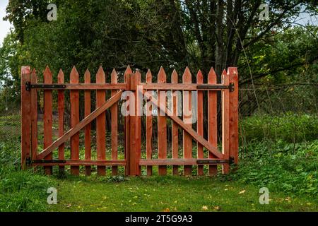 Geschlossene Tore aus hölzernem Pfostenzaun, rot lackiert, vor einem Hintergrund von grünen Bäumen. Stockfoto