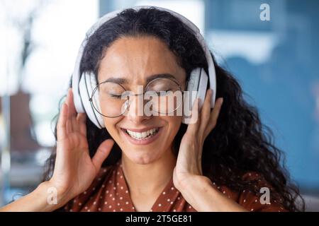 Junge schöne Frau, die am Arbeitsplatz im Büro arbeitet, Nahaufnahme hispanische Frau in Kopfhörern, die fröhlich mitsingt und tanzt, Geschäftsfrau in der Pause ruht, Programmierer glücklich. Stockfoto