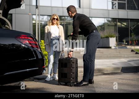 Chauffeur packt einen Koffer in einen Kofferraum, Geschäftsfrau wartet in der Nähe mit einem luxuriösen Taxi während einer Geschäftsreise. Konzept des Unternehmensübertragungskonzepts Stockfoto