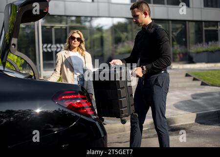 Chauffeur packt einen Koffer in einen Kofferraum, Geschäftsfrau wartet in der Nähe mit einem luxuriösen Taxi während einer Geschäftsreise. Konzept des Unternehmensübertragungskonzepts Stockfoto