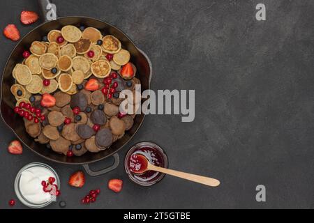 Pfannkuchen und Beeren in der Pfanne. Joghurt und Marmelade auf dem Tisch. Schwarzer Hintergrund. Flache Lagen. Kopierbereich Stockfoto