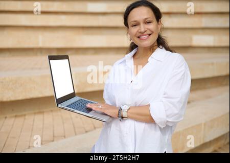 Multi-ethnische angenehm lächelnde Geschäftsfrau, die breit lächelnd in die Kamera blickt, Laptop mit weißem Modell-Digitalbildschirm hält. Werbefläche kopieren Stockfoto