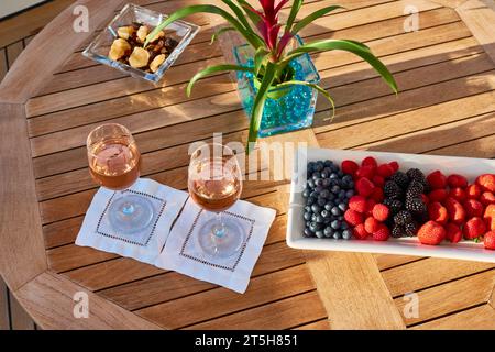 Zwei Gläser Rosenwein auf einem runden Teaktisch mit einem blühenden Kakteen und einer Schüssel Heidelbeeren, Himbeeren und Erdbeeren. Stockfoto