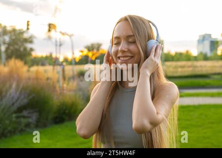Fröhliche junge Frau hört gern Musik und singt im Park. Glückliche Dame in Kopfhörern, die Zeit in der Natur verbringt Stockfoto