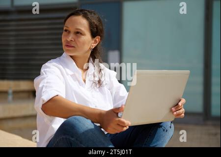 Porträt einer glücklichen jungen lateinamerikanischen Unternehmerin, die auf der Treppe sitzt und draußen einen Laptop benutzt. Online-Schulungen Stockfoto