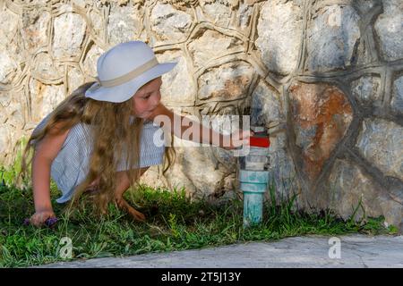 Kind hockt im Sommer an Steinmauer. Lange, lose, blonde Haare. Hut und Sommerkleid Stockfoto