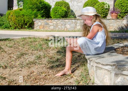 Das Mädchen sitzt barfuß auf einer Steinbank. Weiße Mütze, blaues Kleid, blondes Haar ist locker. Der Fuß eines Beines liegt auf dem Knie des anderen. Stockfoto