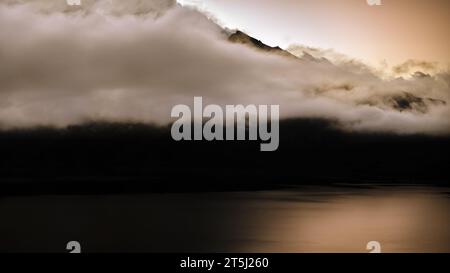 Landschaftsfotos des Lac du Mont Cenis in den französischen Alpen während der goldenen Stunde, dichte Wolken über den Berggipfeln, ruhiges Wasser, dunkel Stockfoto