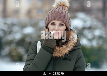 Lächelnde, moderne Frau mittleren Alters im grünen Mantel und braunem Hut draußen im Stadtpark im Winter mit Handschuhen, hygienischem Lippenstift und Mütze. Stockfoto